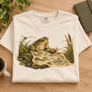 May include: A cream-colored t-shirt featuring a graphic of a green frog wearing glasses, resting its chin on its hand while looking at a map. A snail and a dragonfly are nearby, surrounded by tall grass and cattails. The shirt has a Comfort Colors label.