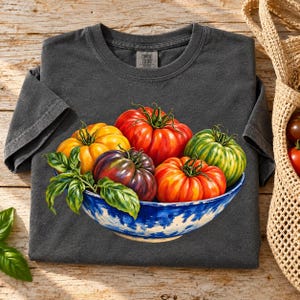 May include: A charcoal gray short-sleeve t-shirt featuring a graphic print of a blue and white bowl filled with various heirloom tomatoes in shades of red, yellow, and purple, accented with green basil leaves. The shirt has a Comfort Colors label.
