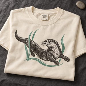 May include: An off-white short-sleeve t-shirt featuring a black ink-style illustration of an otter swimming among green aquatic plants. The inner neck label displays the text Comfort Colors S P CH in a simple black font.