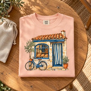 May include: A peach-colored crewneck t-shirt featuring a graphic illustration of a stone bakery with a terracotta roof, blue trim, and a striped curtain. A blue bicycle is parked in front, and the Comfort Colors brand label is visible.