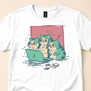 May include: A white short-sleeve t-shirt featuring a graphic illustration of three teal and cream-colored cats with large eyes looking at a laptop screen. The design includes a small cup and a stack of papers, set against a muted red rectangular background.