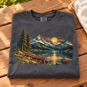 May include: A charcoal gray crew neck t-shirt featuring a graphic print of a red canoe on a rocky lakeshore with pine trees, snow-capped mountains, and a golden sun reflecting on the water. The inner neck label reads Comfort Colors.