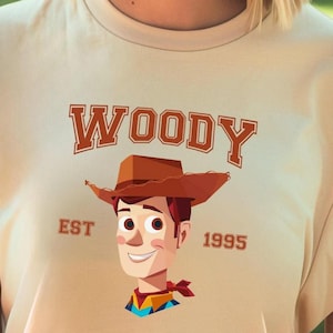 A t-shirt with a cartoon image of a smiling character wearing a cowboy hat and the text "WOODY EST 1995" printed on it.