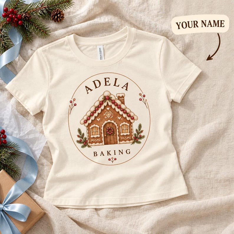 Cream gingerbread house tee laid flat beside blue ribbon and greenery.