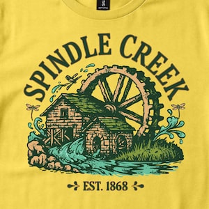 May include: A yellow t-shirt featuring a graphic of a water mill with the text "SPINDLE CREEK" arched above and "EST. 1868" below. The illustration includes a wooden building, a large water wheel, splashing water, and dragonflies in shades of green, brown, and teal.
