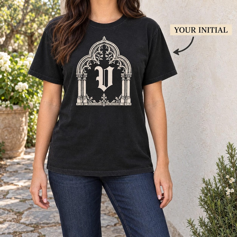 Woman in a black tee with a cream trefoil gothic arch framing a blackletter V initial