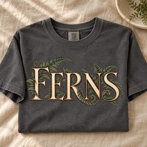 May include: A charcoal gray short-sleeve t-shirt featuring the word FERNS in large, cream-colored serif lettering. The text is intertwined with detailed green fern leaf illustrations. The shirt has a crew neckline and a Comfort Colors label.