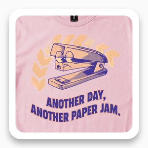 May include: A light pink t-shirt featuring a graphic of a cartoon stapler with a tired expression. Behind the stapler is a yellow chevron pattern. Below the graphic, the text reads: ANOTHER DAY, ANOTHER PAPER JAM. The shirt has a crew neckline.
