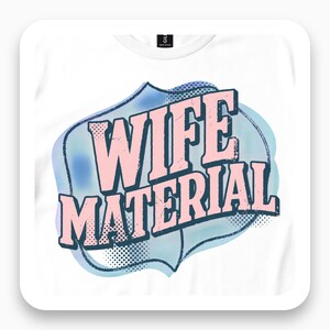 May include: A white t-shirt featuring a graphic design with the text WIFE MATERIAL in large, light pink, block-style letters. The text is outlined in dark blue and set against a soft, light blue, cloud-like shape with a subtle black dot pattern.