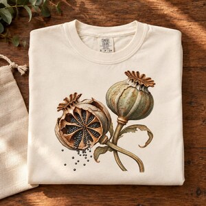May include: A cream-colored t-shirt featuring a graphic print of two poppy seed pods, one intact and one open with visible seeds spilling out. The shirt has a Comfort Colors brand label at the inner neckline, indicating a size S-CH.