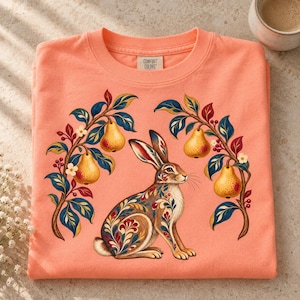 May include: A peach-colored crew neck t-shirt featuring a central graphic of a rabbit with floral patterns on its fur, flanked by two branches laden with yellow pears and small white flowers. The inner neck label reads Comfort Colors.