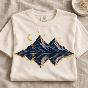May include: A cream-colored short-sleeve t-shirt featuring a graphic of dark blue mountains with a golden crescent moon and stars above, reflecting in water below. The inner neck label displays the text Comfort Colors S P CH.