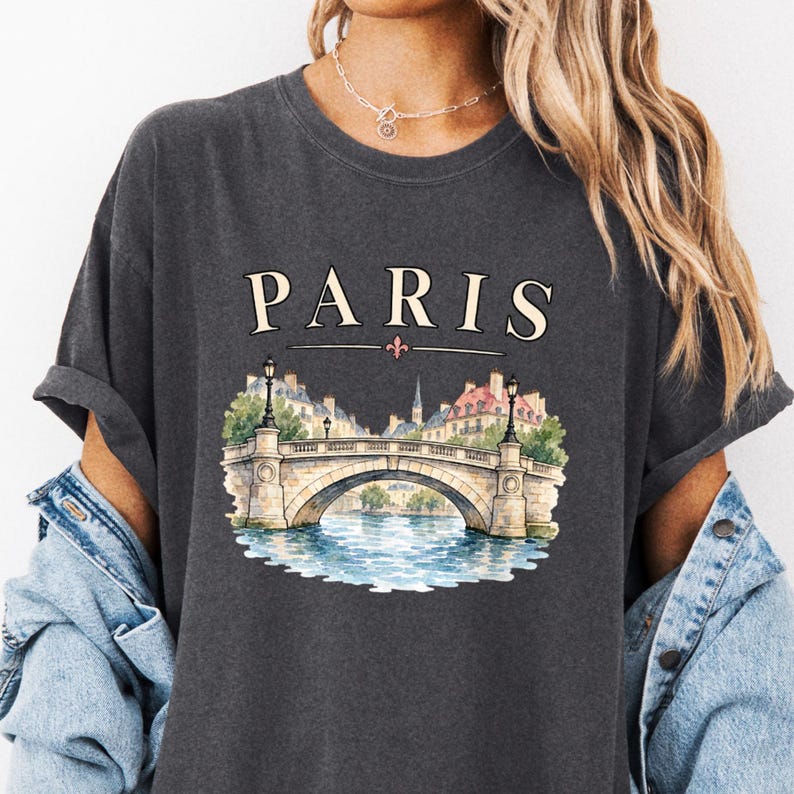 Charcoal tee close-up on a model with Paris lettering above a stone bridge and blue river.