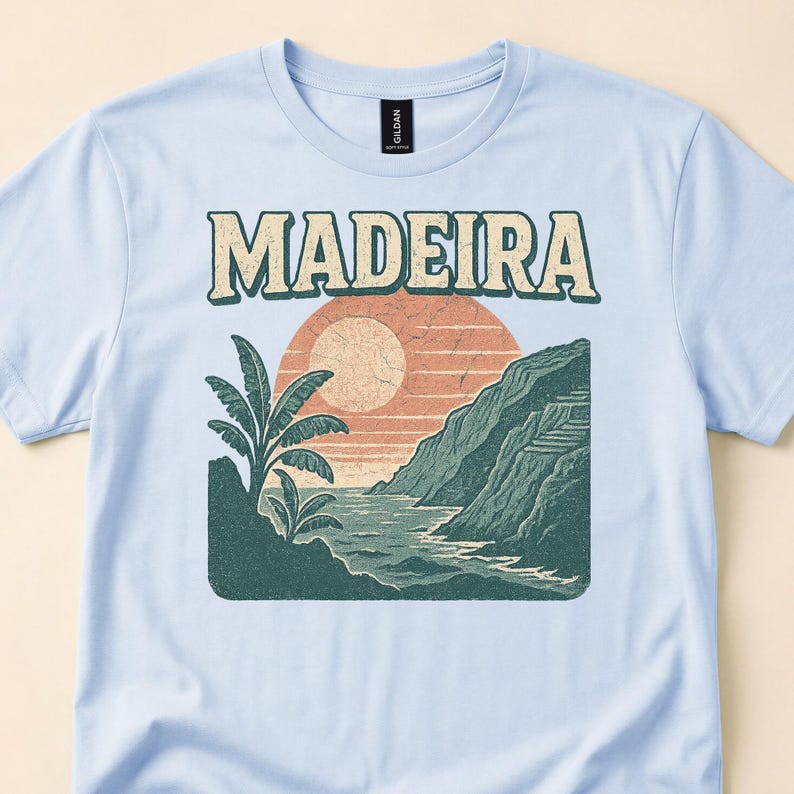 Pale blue tee close-up with Madeira lettering above green coastal cliffs and a peach sunset.