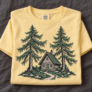 May include: A pale yellow short-sleeve t-shirt featuring a graphic of a small cabin nestled between two tall pine trees. The cabin has a green door and roof, and a stone path leads to the entrance. The Comfort Colors brand label is visible at the neck.