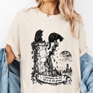 May include: A beige short-sleeved t-shirt featuring a black ink-style graphic of two cats on a crumbling stone pillar. The design includes a moonlit landscape and a banner with the text: i contemplate the fall fall is my only ground.
