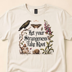 May include: A cream-colored short-sleeve t-shirt featuring a graphic print of a black crow, a moth, a snail, mushrooms, and purple flowers. The text "Let your Strangeness Take Root" is printed in black in the center of the botanical illustration.