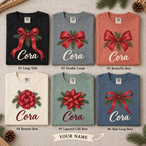 May include: Six t-shirts in black, gray, terracotta, cream, sage green, and blue, each featuring a unique red holiday bow with pine needles and the name Cora printed in white or dark red script. Each shirt has a Comfort Colors size L label.