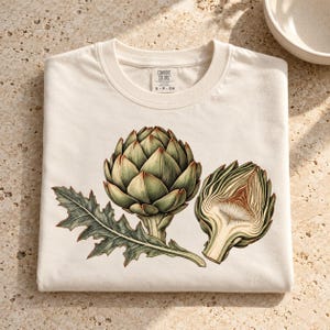 May include: A cream-colored crew neck t-shirt featuring a detailed botanical illustration of a whole green artichoke and a halved artichoke with a jagged leaf. The inner neck label displays the text Comfort Colors S-P-CH.