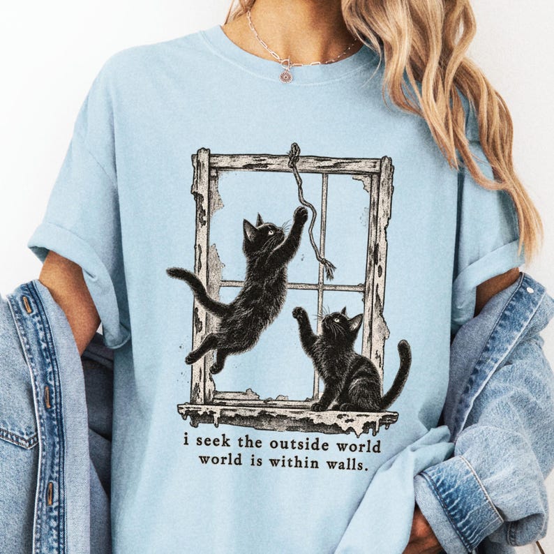 Pale blue tee close-up on a model showing two cats at a window and I Seek the Outside World text.