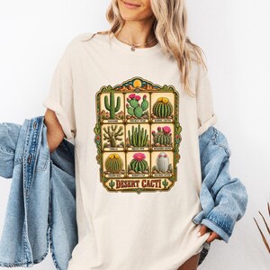 May include: A cream-colored t-shirt featuring a graphic print of nine different cacti arranged in a grid. Each cactus is labeled with its name, including Saguaro, Prickly Pear, and Barrel Cactus. The text at the bottom reads Desert Cacti.