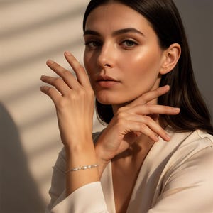 Könnte beinhalten: Ein filigranes silbernes Armband mit kleinen, gleichmäßig verteilten weißen Perlen. Das Armband wird am Handgelenk getragen, mit den Händen und dem Gesicht der Person im Bild. Der Hintergrund ist eine weiche, neutrale Farbe mit Schatten.