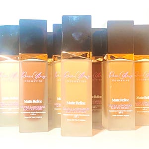 May include: Several bottles of Rose Glow Cosmetics liquid foundation. The bottles are rectangular with a matte finish and dark caps. The text "Matte Refine" and "Ultra Longwear Matte Finish" is printed on the bottles.