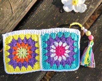 May include: A colorful, crocheted pouch featuring two square panels with floral designs in yellow, orange, purple, and turquoise. The pouch has a white border and a tassel with beads. The background is a weathered wooden surface.