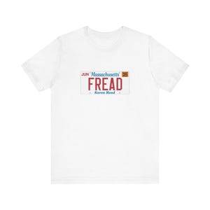 May include: White t-shirt featuring a Massachusetts license plate design. The license plate reads "FREAD" in red letters, with "Massachusetts" and "JUN 25" at the top. The text "Karen Read" is at the bottom. A casual, everyday wear tee.