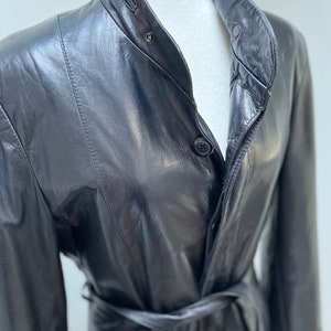 Puede incluir: Una gabardina de cuero negro con cinturón en la cintura y cierre de botones. La gabardina tiene un cuello alto y mangas largas.