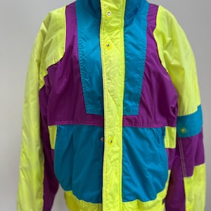 Può includere: Una giacca da sci vintage con un design a blocchi di colore giallo brillante, viola e turchese. La giacca ha un colletto alto e una chiusura con cerniera.