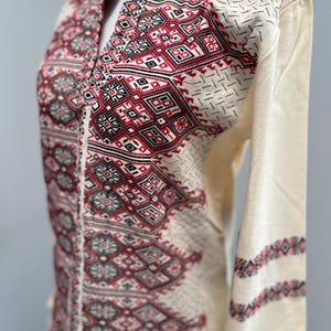 May include: A white long-sleeved shirt with a geometric pattern in red, black, and gray. The pattern is embroidered on the front of the shirt.