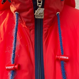 Peut inclure: Veste rouge à capuche avec une fermeture éclair bleu marine et la marque K-Way sur le tirette de la fermeture éclair et le bas de la veste.