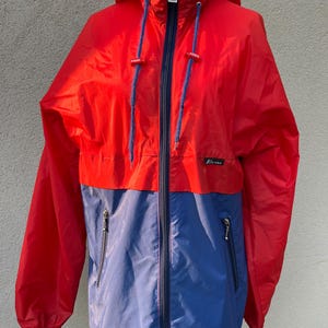 May include: A red and blue hooded jacket with a zipper closure and two pockets. The jacket has a logo on the left side of the chest.