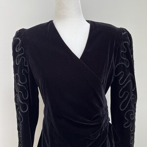 May include: Black velvet wrap top with long sleeves. The top features decorative embroidered detailing on the sleeves and a gathered waist. The neckline is a V-shape. The top is displayed on a mannequin.