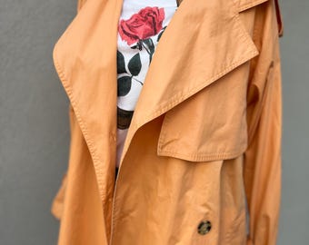 May include: A long, peach-colored trench coat with a large collar and a double-breasted closure. The coat has a belt at the waist and buttons on the front. The coat is worn over a white t-shirt with a floral print and the text "CITY AND COLOUR".