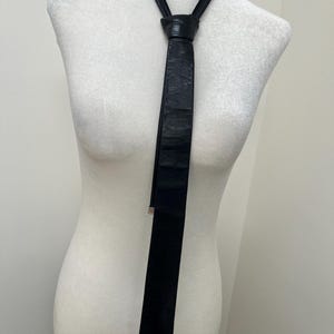 Pode incluir: Uma gravata preta com acabamento texturizado semelhante a couro, amarrada ao redor do pescoço de um manequim branco. A gravata apresenta um design fino e alongado com uma ponta afiada e um pequeno detalhe metálico na extremidade da tira.