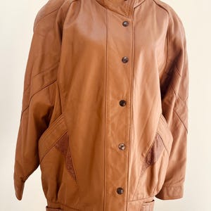 May include: A tan leather jacket with a high collar and button closure. The jacket features textured panels on the shoulders and pockets. It has a ribbed hem and cuffs. The jacket is displayed on a mannequin.