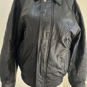 May include: Black leather jacket with a collared neckline, two zipped pockets, and a button closure. The jacket has a bomber-style design with ribbed cuffs and a hem. The leather appears to be slightly textured.