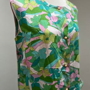 May include: Sleeveless floral print top in shades of green, blue, pink, and white. The top features a round neckline, wooden buttons, and two front pockets. The fabric has a silky sheen, and the floral pattern is a repeating design.