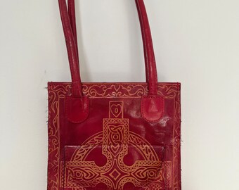 May include: A red leather tote bag with a Celtic cross design in gold. The bag has two top handles and a small pocket on the front.