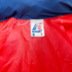 May include: A red and blue jacket with a white K-Way label. The label has the text "K-WAY" and "S" in black lettering. The number "7" is printed in black on the label.
