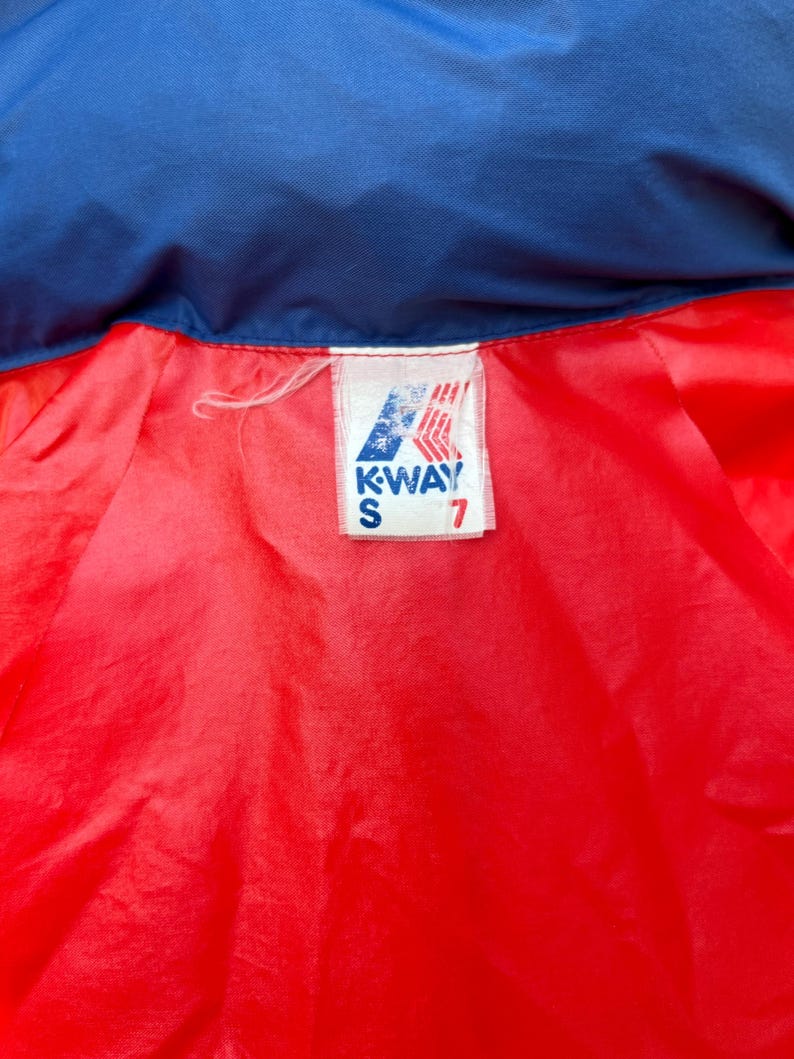 May include: A red and blue jacket with a white K-Way label. The label has the text "K-WAY" and "S" in black lettering. The number "7" is printed in black on the label.
