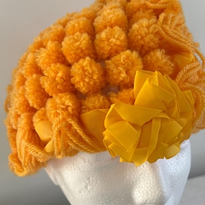 Puede incluir: Un gorro de crochet amarillo brillante con un gran diseño de pompones y un adorno de flor de tela amarilla.