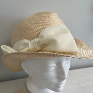 Puede incluir: Un sombrero de paja beige con un gran lazo de satén blanco. El sombrero tiene un ala ancha y está adornado con una cinta de satén blanco. El sombrero está sobre una cabeza de maniquí blanca. El sombrero tiene una etiqueta que dice "Jacqueline".