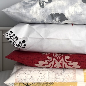 May include: A stack of four decorative cushions with different patterns. The top cushion is grey with a floral design, the second is white with black floral accents, the third is red with a pink floral design, and the bottom cushion is yellow with a handwritten script and a black floral design.