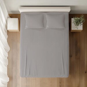 May include: An overhead shot of a bed with grey bedsheets and two matching pillows. The bed is against a white headboard. Two small white bedside tables flank the bed, one with a plant. The floor is wooden.