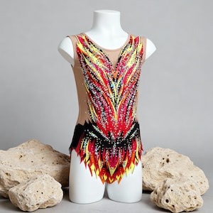 May include: A sleeveless gymnastics leotard on a white mannequin. The leotard features a vibrant design with red, yellow, and black accents, embellished with sparkling rhinestones. The garment is displayed against a neutral background with decorative stones.