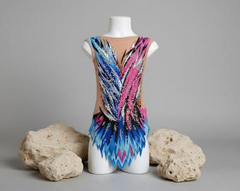 Puede incluir: Maillot sin mangas para gimnasia rítmica, expuesto sobre un maniquí blanco. El maillot presenta una base de malla beige con un diseño vibrante de patrones en forma de pluma azul, rosa y blanco. El diseño está acentuado con purpurina plateada.