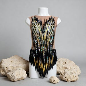 May include: A sleeveless gymnastics leotard on a white mannequin. The leotard features a nude-colored bodice with a black and gold design, embellished with iridescent sequins. The bottom of the leotard has a black, jagged edge.